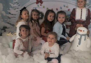 Grupa dziewczynek pozuje do zdjęcia z bałwankiem, w zimowo- świątecznej scenerii.